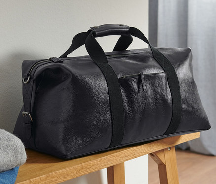 Eine schwarze Leder-Weekender liegt auf einer Holzbank neben gefalteter Kleidung.