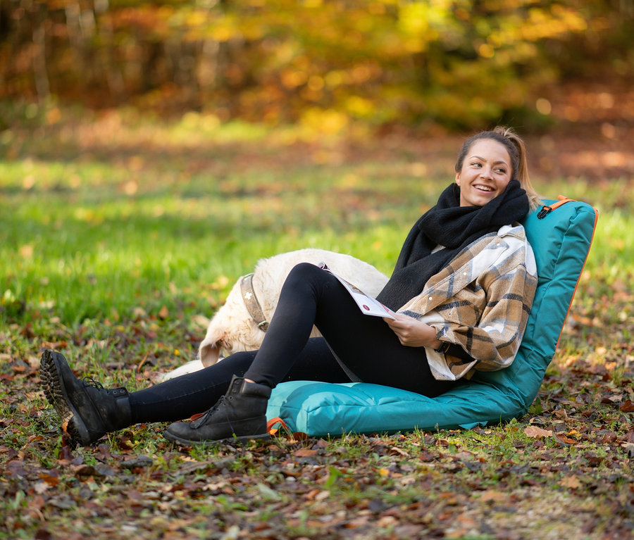 Frau sitzt mit Hund auf einer tragbaren Sitzmatte im Freien und liest.