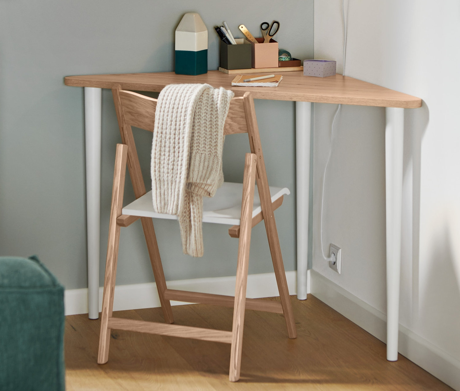 Ein Klappstuhl aus FSC®-zertifiziertem Eschenholz steht an einem dreieckigen Tisch. An der Wand hängt eine weiße Wandleuchte aus Metall.