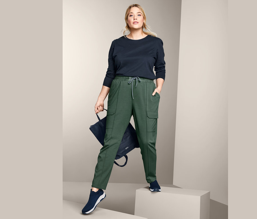 Frau posiert in grüner Joggpants mit Cargo-Taschen und schwarzem Langarmshirt.