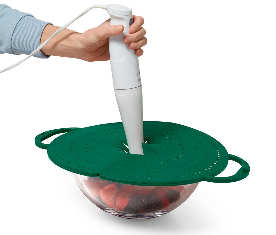 Eine Hand hält einen Stabmixer über eine Schüssel mit Früchten, die mit einem grünen 4-in-1-Pfannen-Spritzschutz abgedeckt ist.