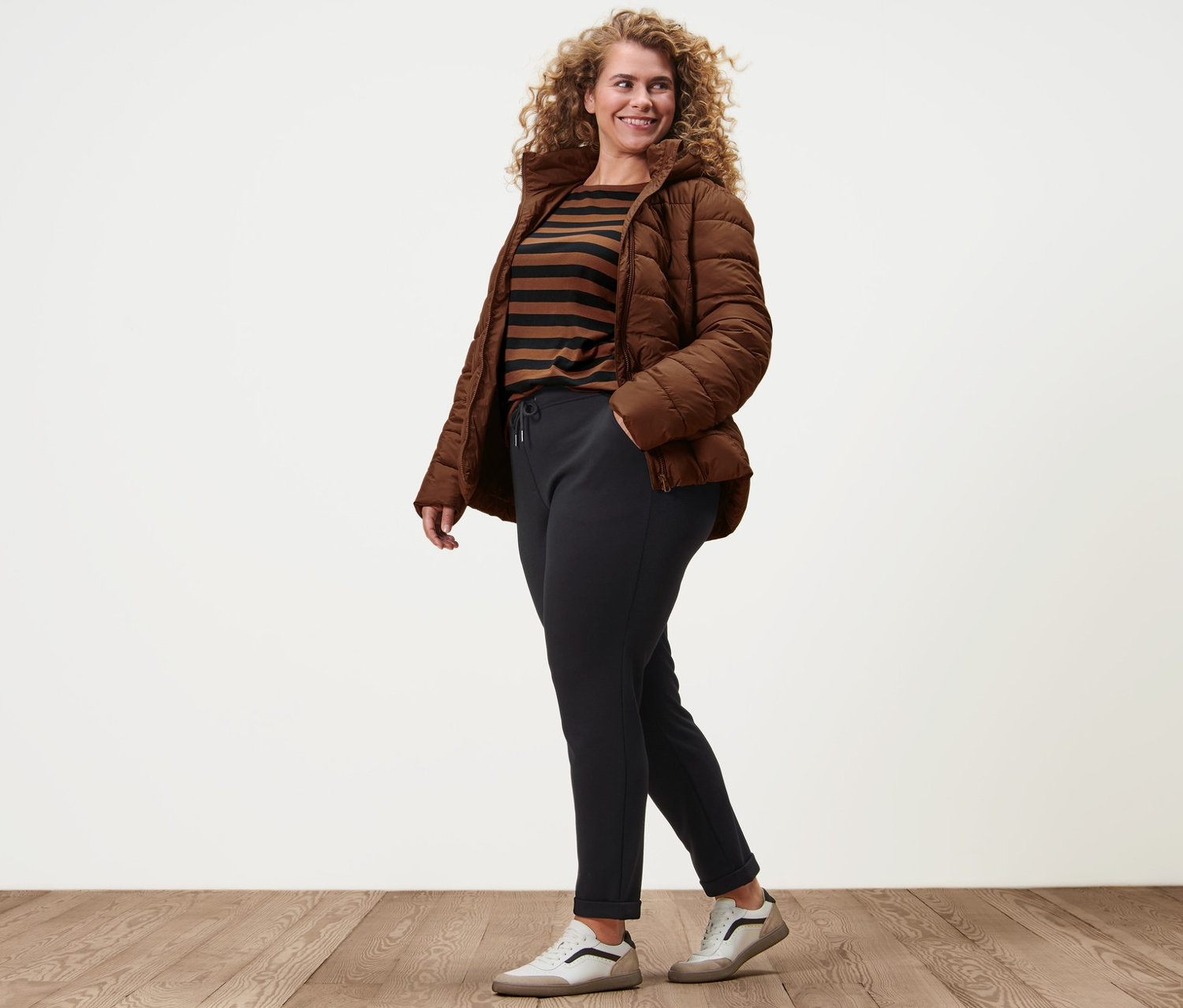 Eine Frau geht und trägt eine cognacfarbene Steppjacke mit Kapuze, ein gestreiftes Shirt mit 3/4-Arm, Joggpants und Leder-Sneaker.