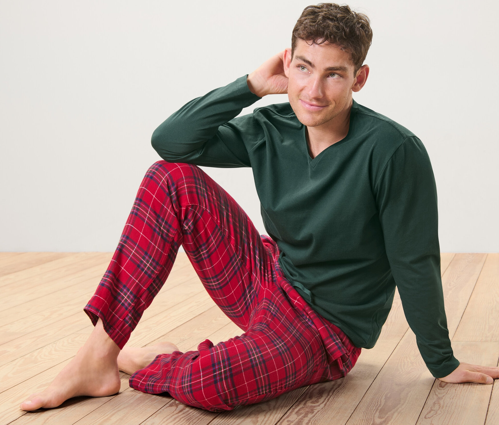 Mann sitzt auf Holzboden und trägt einen grünen Pyjama mit rot karierter Flanellhose.