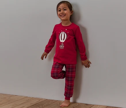 Lächelndes Mädchen steht an einer Wand, trägt einen roten Pyjama mit karierter Hose und einem Ballon-Aufdruck, der im Dunkeln leuchtet.
