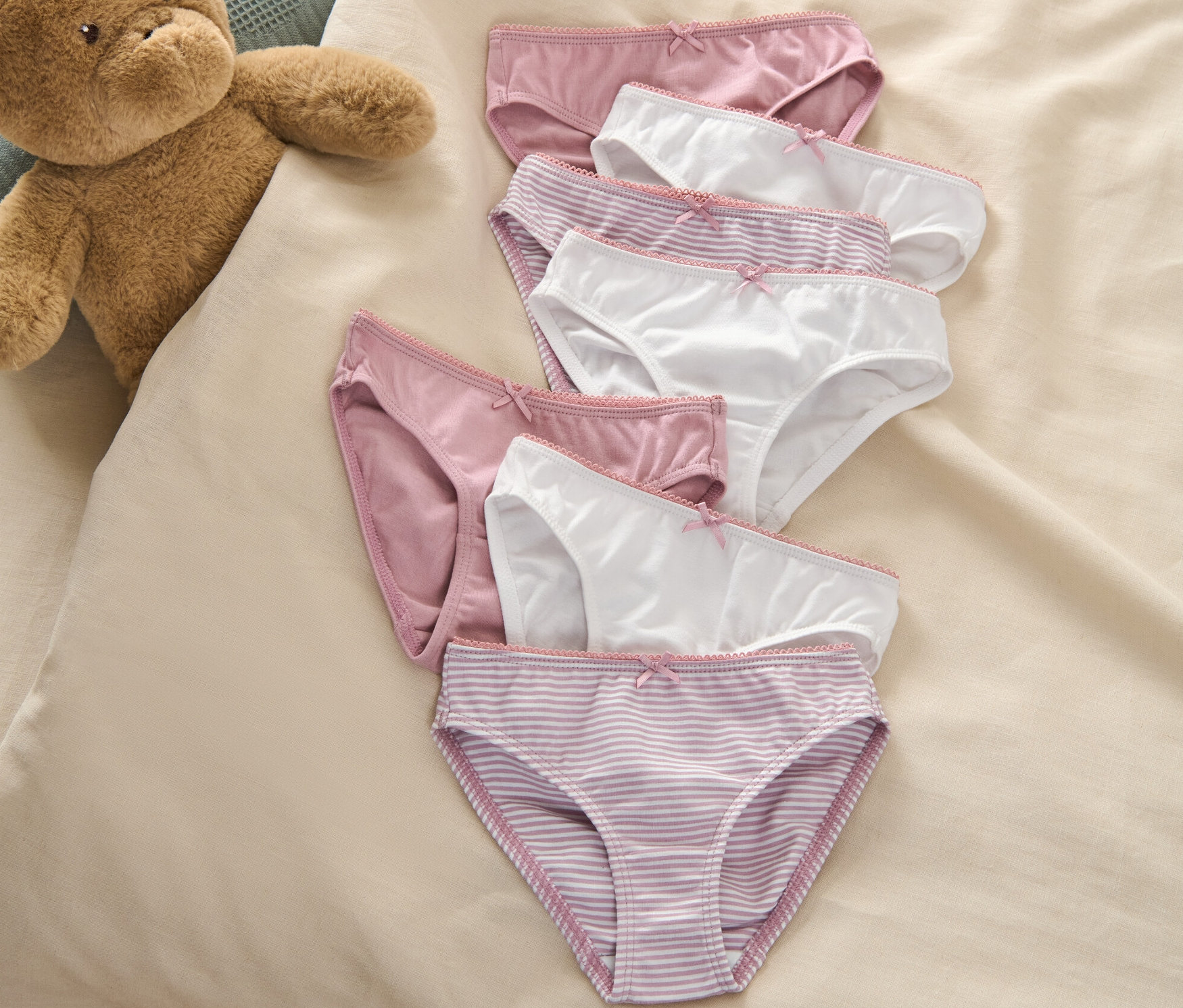 Sieben Kinder-Slips liegen neben einem Teddybär auf einem Bett.