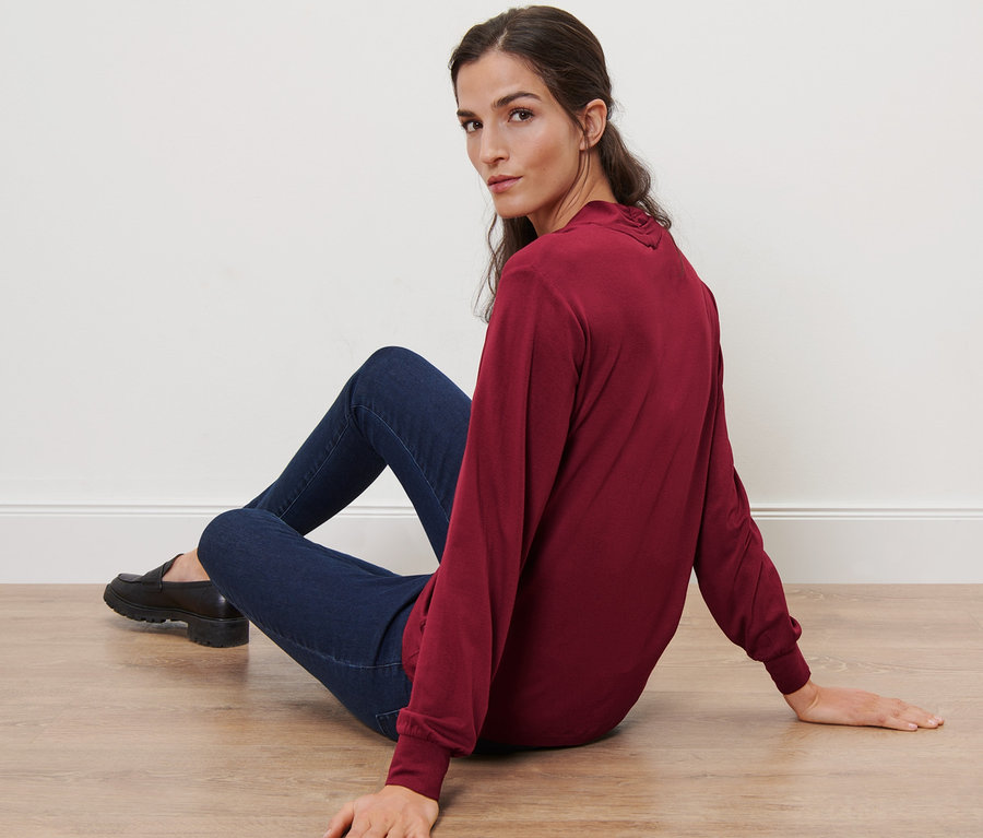 Frau sitzt auf dem Boden und trägt ein bordeauxrotes Langarmshirt mit V-Ausschnitt.