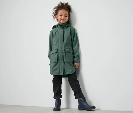Mädchen steht an Wand angelehnt in Kinder-Softshellmantel, schwarzen Hosen und blauen Schuhen.