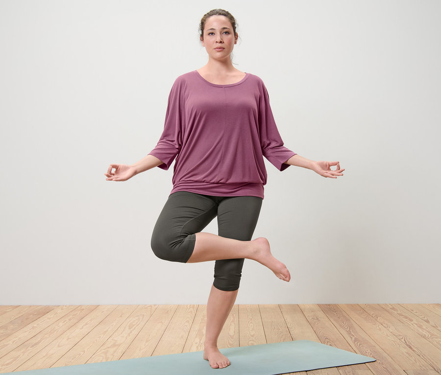 Frau steht in der Baum-Pose auf einer Yogamatte. Sie trägt ein Yogashirt in Mauve und eine 3/4-Sporttight in Oliv.