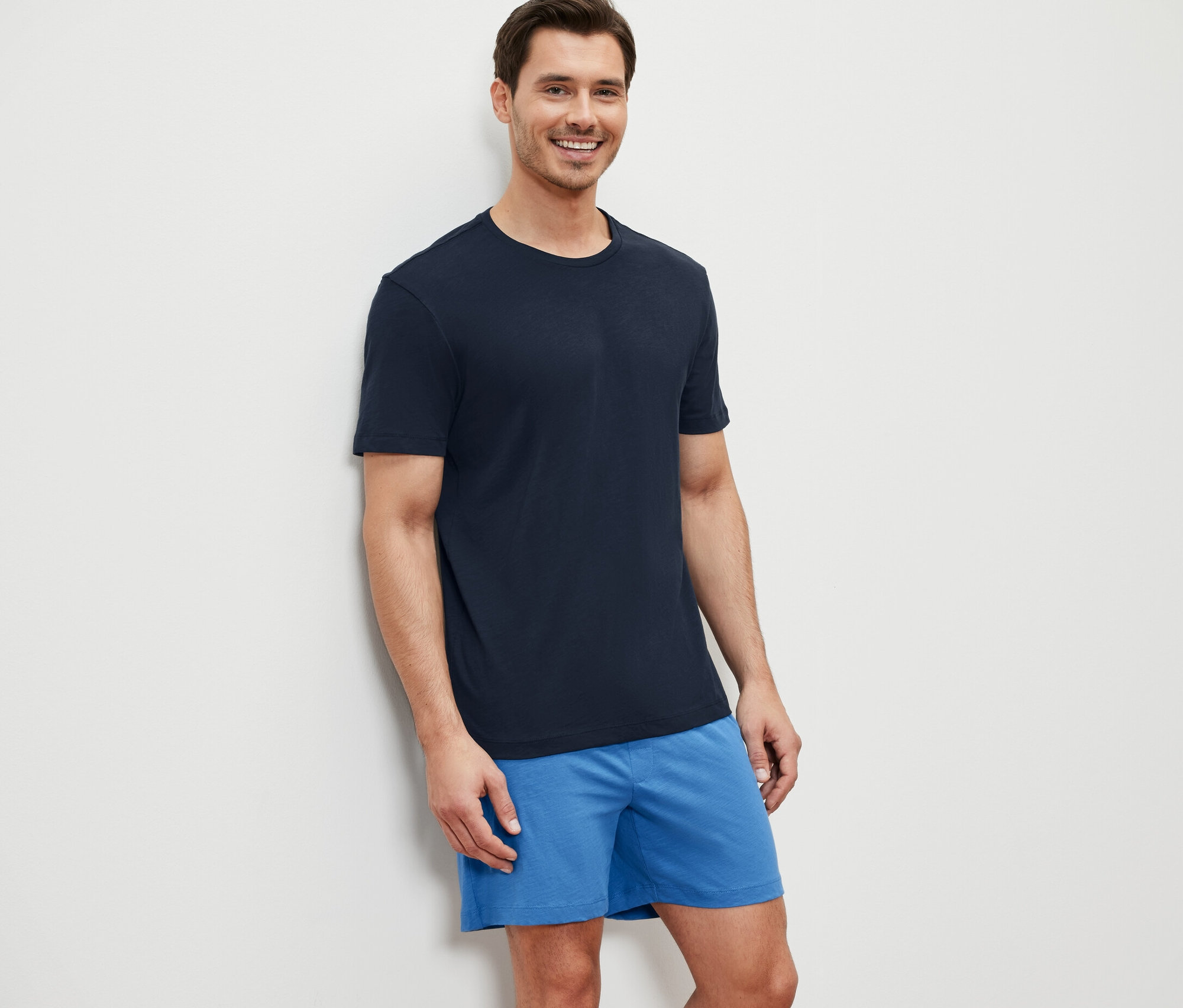 Ein Mann lächelt, an eine Wand gelehnt, in einem Shorty-Pyjama.