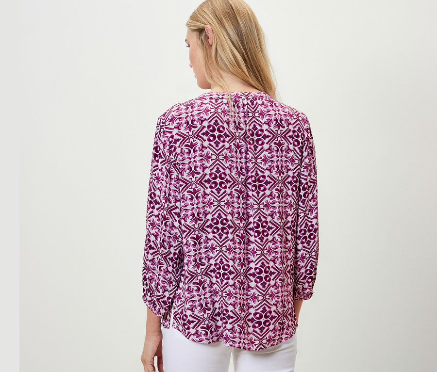 Rückansicht einer Frau mit bedruckter Tunika-Bluse, berry, und weißen Hosen.