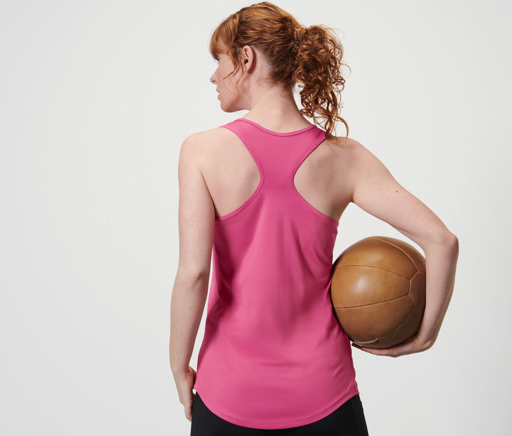 Frau hält von hinten einen braunen Ball in einem pinken Funktionstop von einem zweiteiligen Set und schwarzen Leggings.