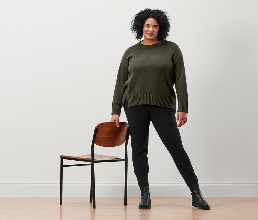 Frau posiert neben einem Stuhl und trägt eine schwarze Stretchhose und einen Strickpullover mit Saumschlitz.