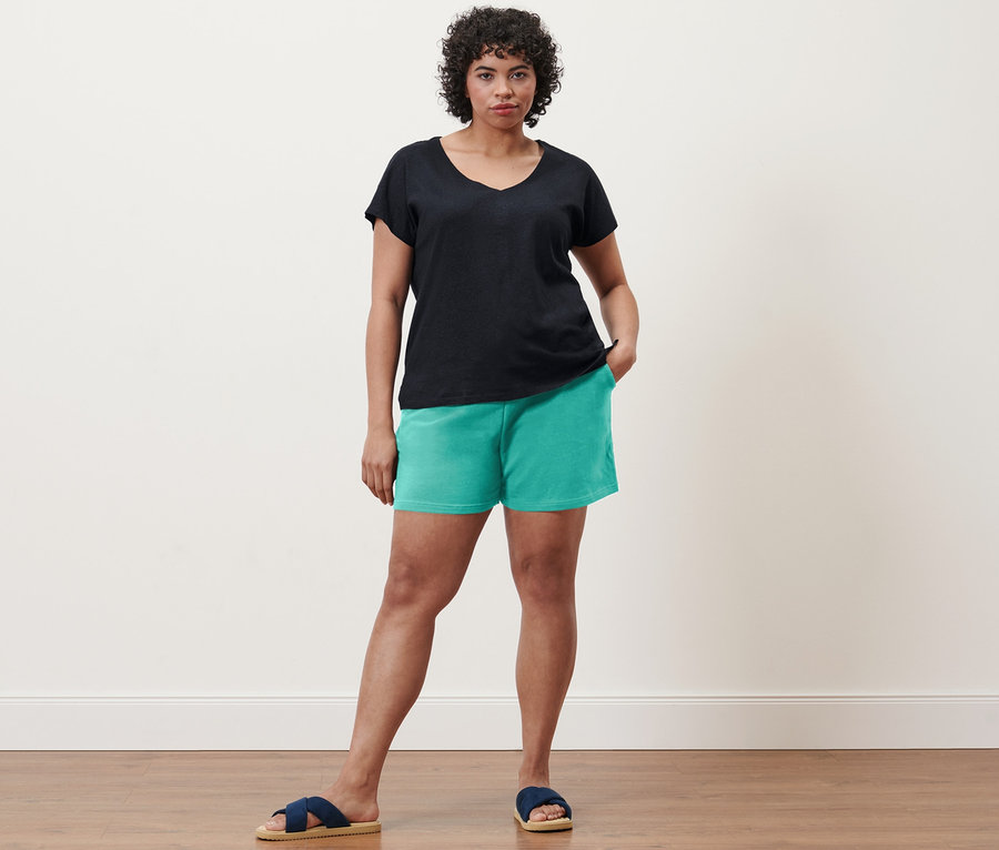 Eine Frau steht und trägt ein dunkelblaues Shirt aus Leinenmix, aqua Sweatshorts und Komfortsandalen.