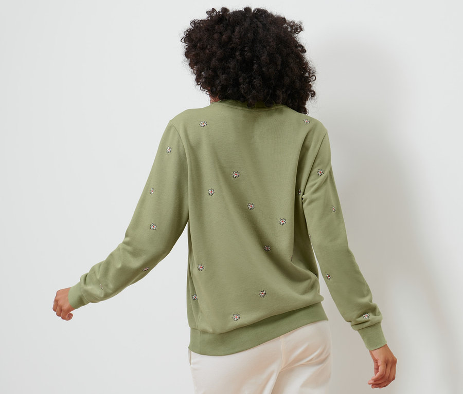 Rückansicht einer Frau mit dunklen lockigen Haaren, die ein besticktes Sweatshirt in Grün und helle Hosen trägt.
