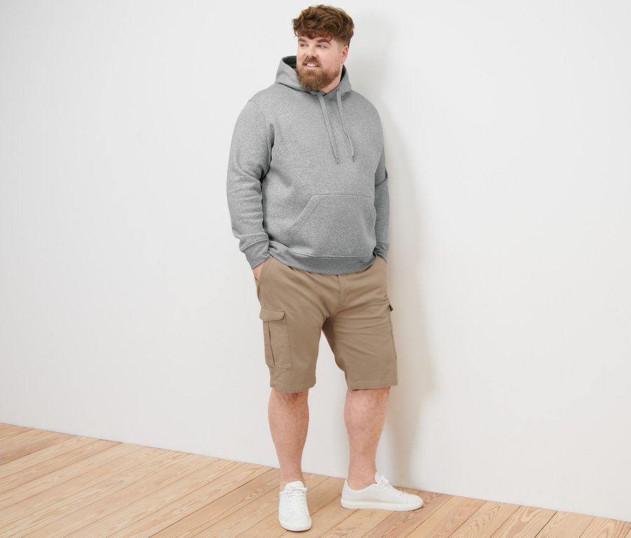 Mann mit Bart, der einen grau melierten Hoodie und eine beige Cargo-Shorts trägt und steht.