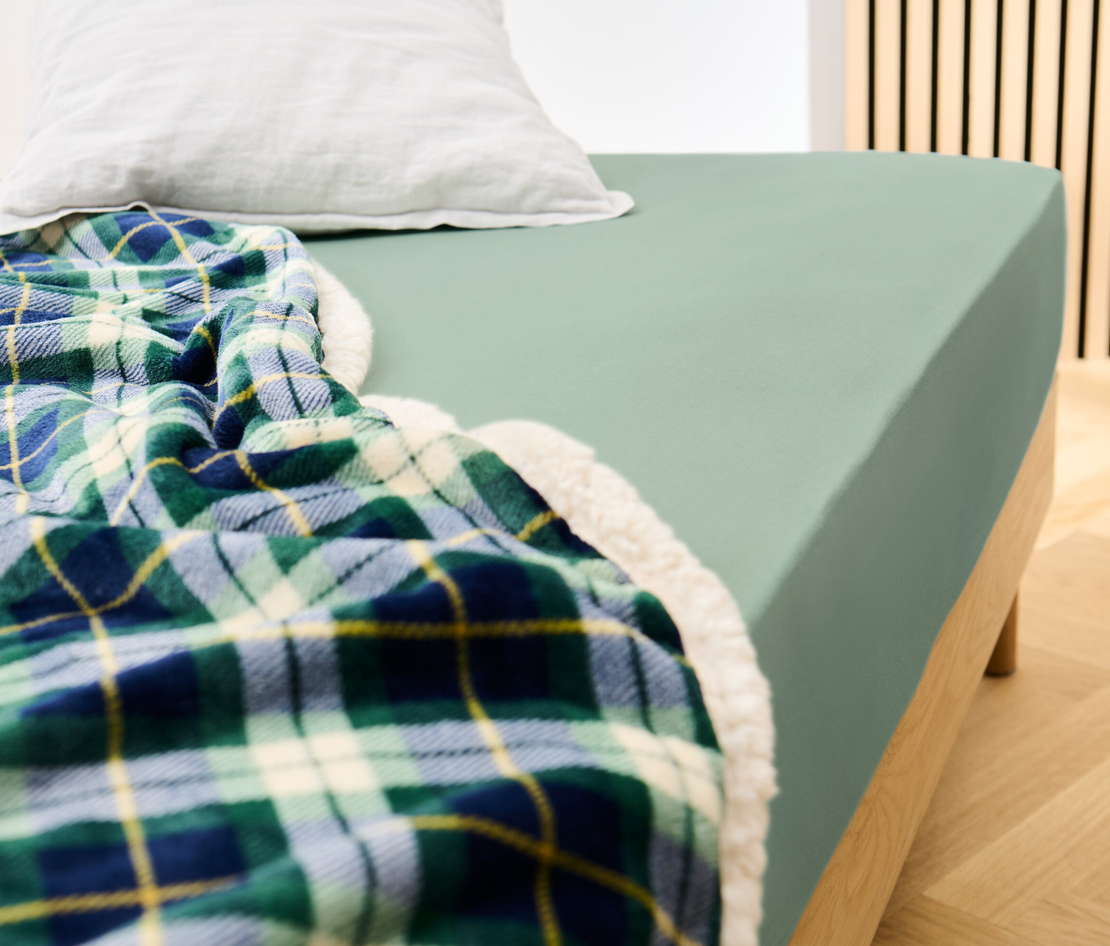 Bett mit einem salbeifarbenen Jersey-Spannbettlaken und einer karierten Decke.