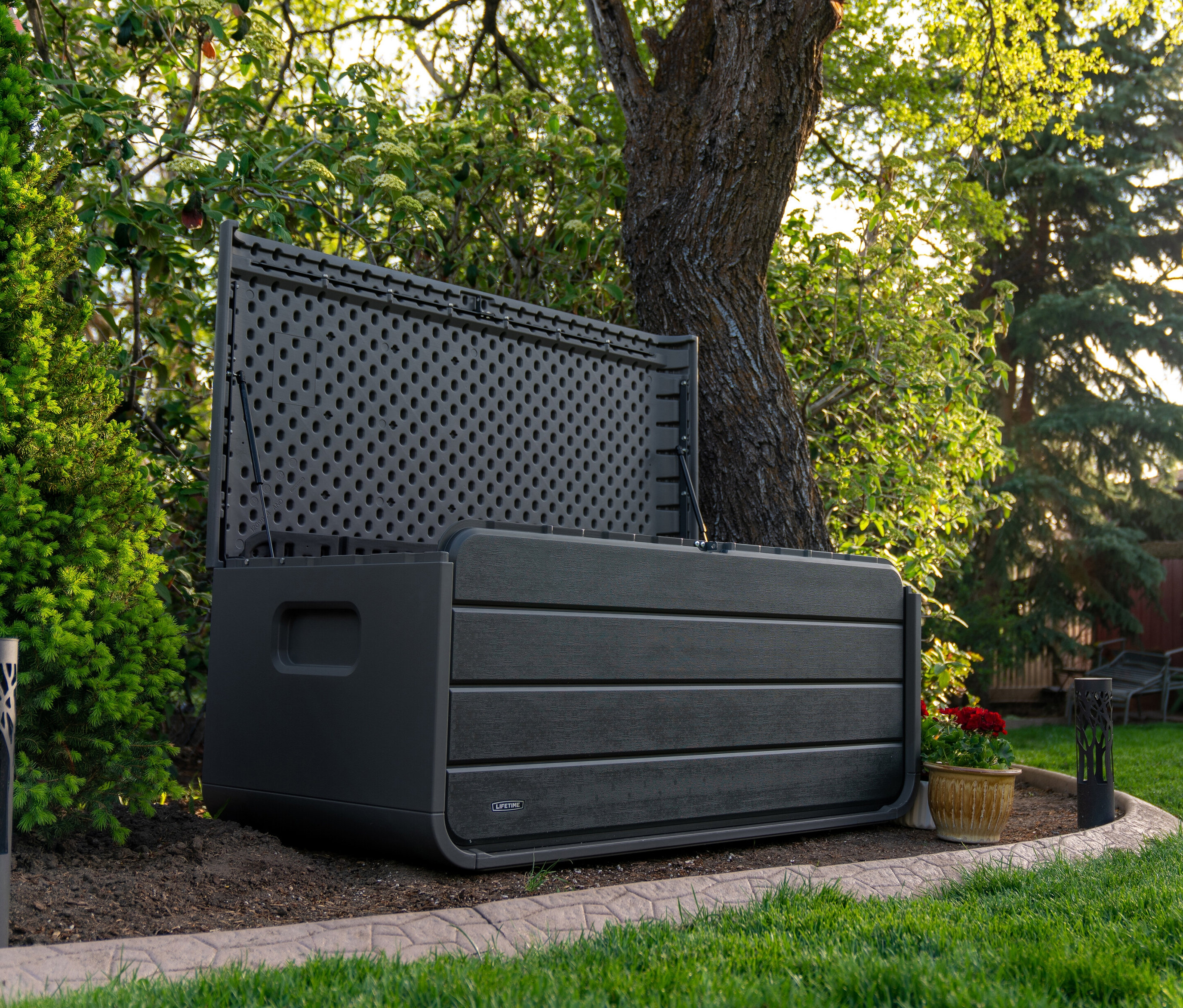 Eine graue Aufbewahrungsbox mit 515 l Fassungsvermögen steht im Freien neben einem Baum, der Deckel ist offen.