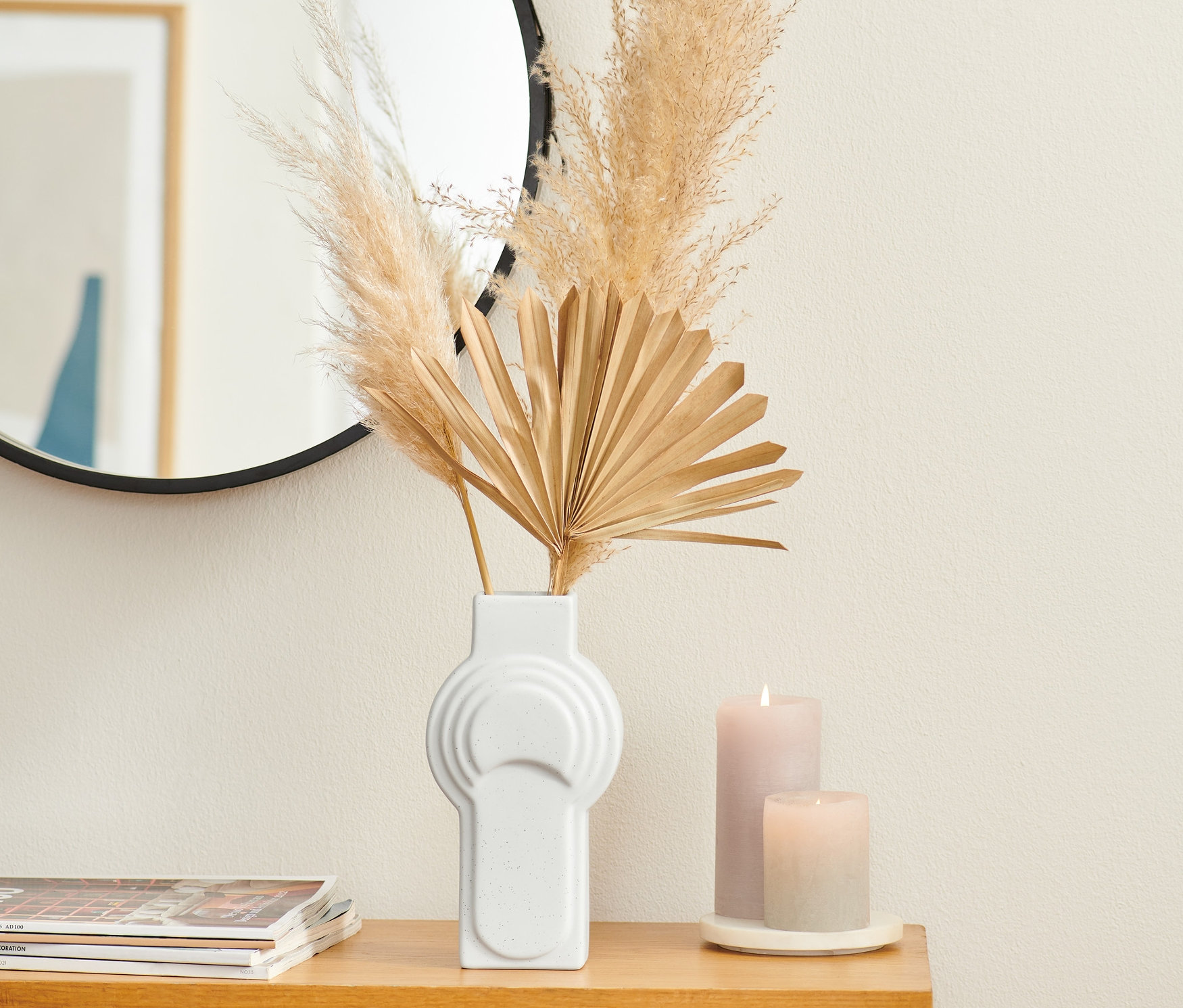 Auf einem Holztisch stehen eine große Vase aus Keramik mit Pampasgras und getrockneten Palmen, daneben zwei Kerzen und im Hintergrund ein Spiegel.