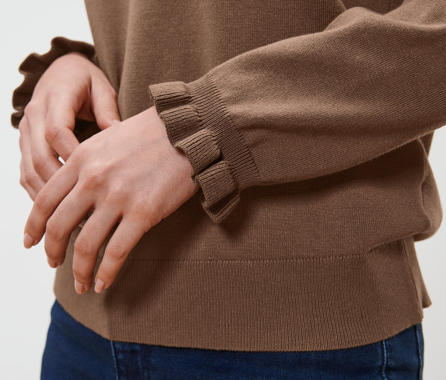 Person trägt einen braunen Feinstrickpullover mit Rüschendetail und blaue Jeans.