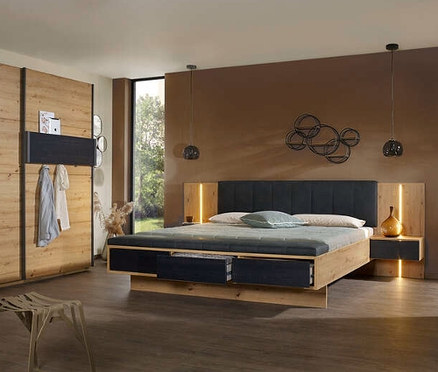 Schlafzimmer mit Holzbett und Schrank. Auf dem Bett liegen Kissen und die Nachttische sind beleuchtet. Rauch Bettanlage "Apollon", ca. 160 x 200 cm und 180 x 200 cm, in Eichenoptik sichtbar.