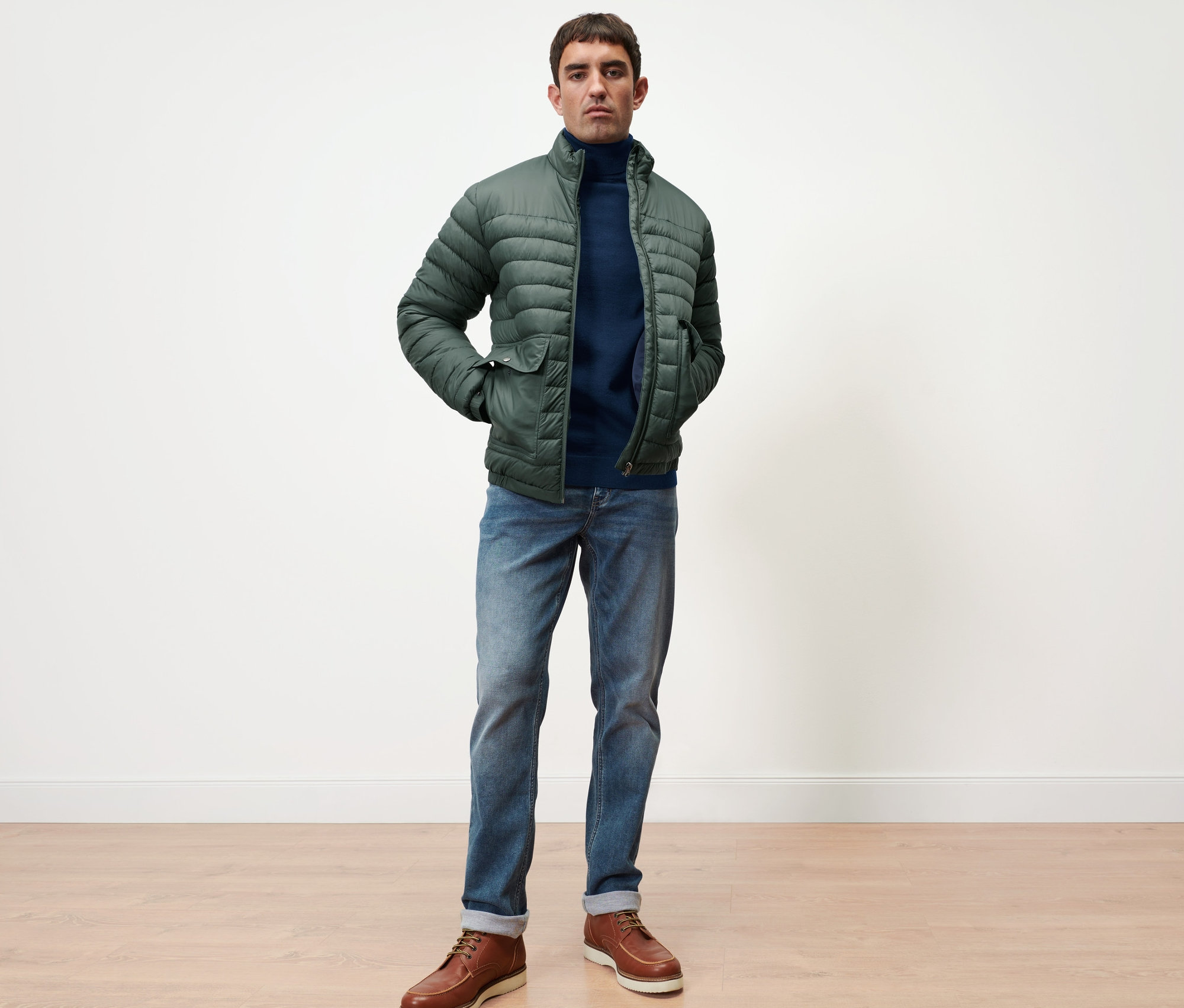 Mann steht, trägt einen navy Merino-Pullover und eine grüne Steppjacke.