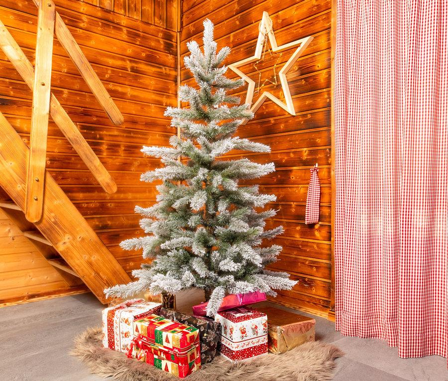 In der Ecke eines Holzraums steht eine schneebedeckte Cedar Fort Kiefer mit Metallständer, 150 cm, umgeben von Geschenken.