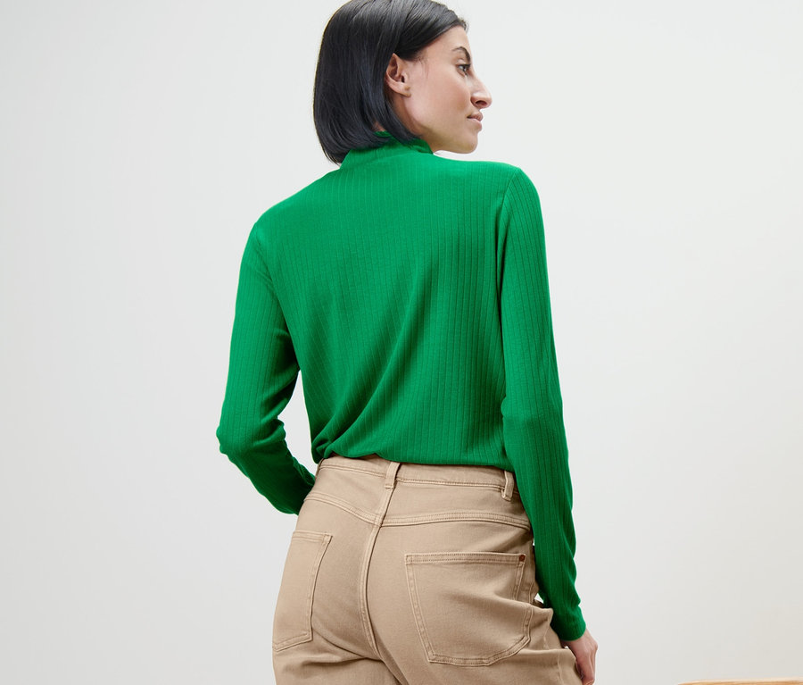 Frau mit grünem Langarmshirt steht mit dem Rücken zur Kamera neben einem Stuhl.