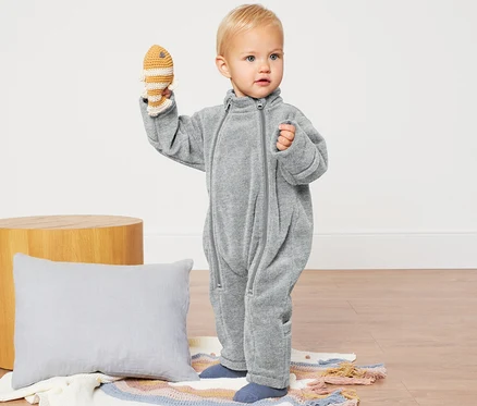 Kleinkind hält gehäkelten Fisch und trägt einen grauen Baby-Fleece-Einteiler.