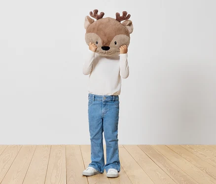 Ein Kind hält ein Stofftier-Rentier vor sein Gesicht, steht auf einem Holzboden vor einer weißen Wand.