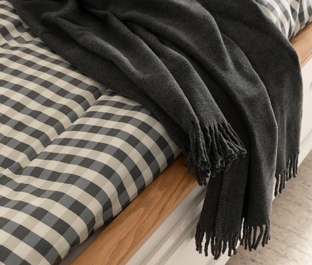 Dunkelgraue SCHÖNER WOHNEN-Kollektion Wohndecke »BASICS« liegt auf einem Bett mit grau-weiß karierter Bettwäsche.
