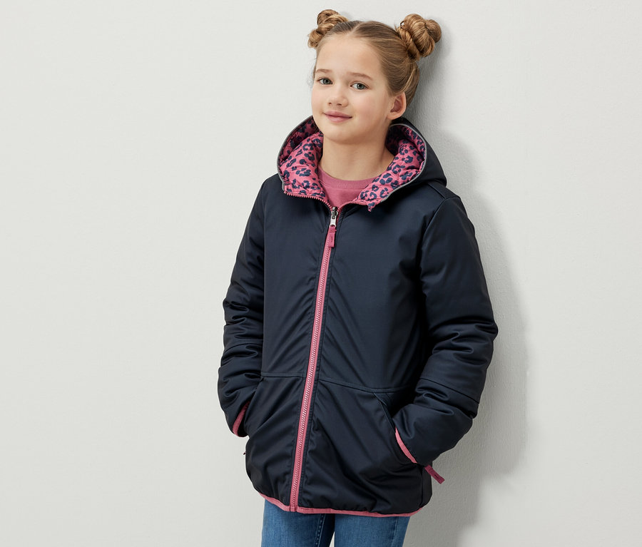 Mädchen posiert in 2-in-1 Kinder Softshell- und Steppjacke mit Kapuze, pinkem Reißverschluss und Bündchen.