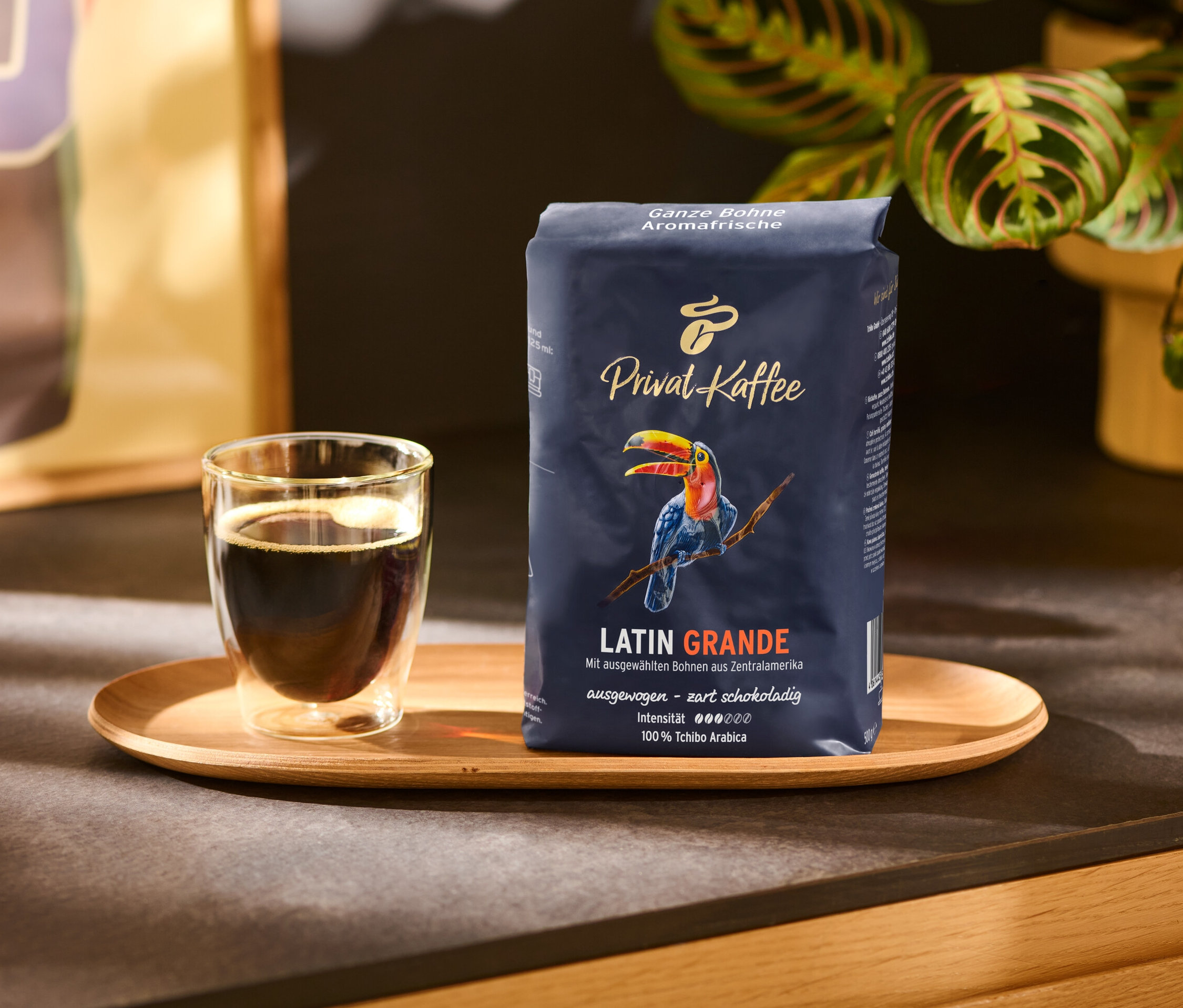 Ein Glas Kaffee und eine Packung Privat Kaffee Latin Grande - 500g Ganze Bohne auf einem Holztablett.