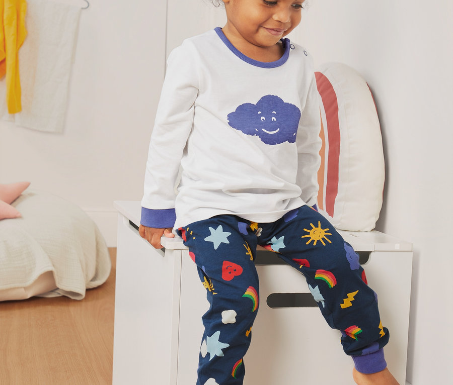 Mädchen sitzt auf einer weißen Truhe und trägt 2 Pyjamas aus Bio-Baumwolle. Das Oberteil ist weiß mit einer blauen Wolke, die Hose ist dunkelblau mit Mustern.