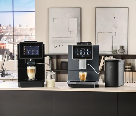 Zwei Kaffeemaschinen mit Touchscreens bereiten Kaffee in Gläsern zu. Im Hintergrund sind ein Fenster und dekorative Bilder zu sehen.