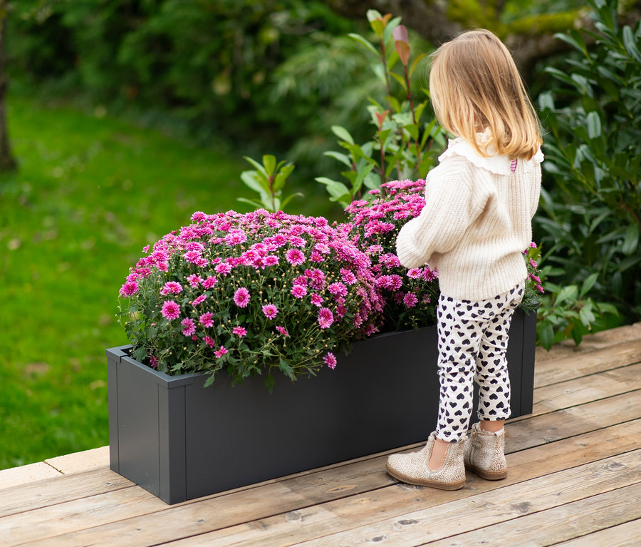 Mädchen steht neben einem grauen Pflanzkasten mit rosa Blumen.