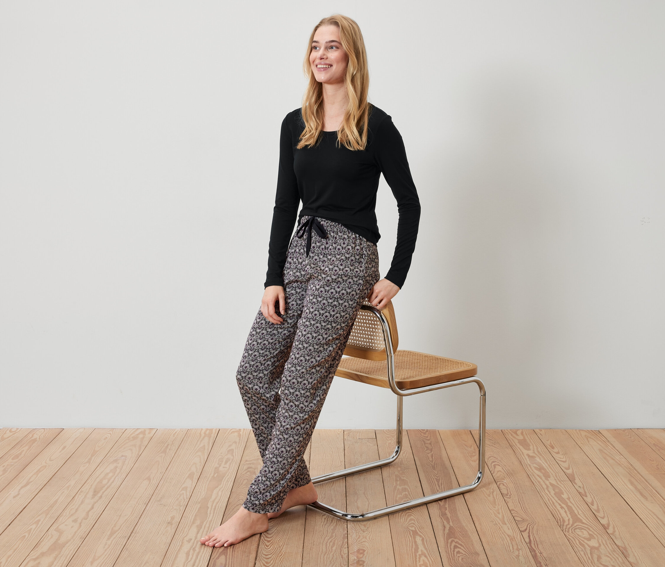 Blonde Frau in schwarzem Oberteil und gemusterter Hose lehnt an einem Stuhl.