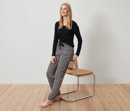 Blonde Frau in schwarzem Oberteil und gemusterter Hose lehnt an einem Stuhl.