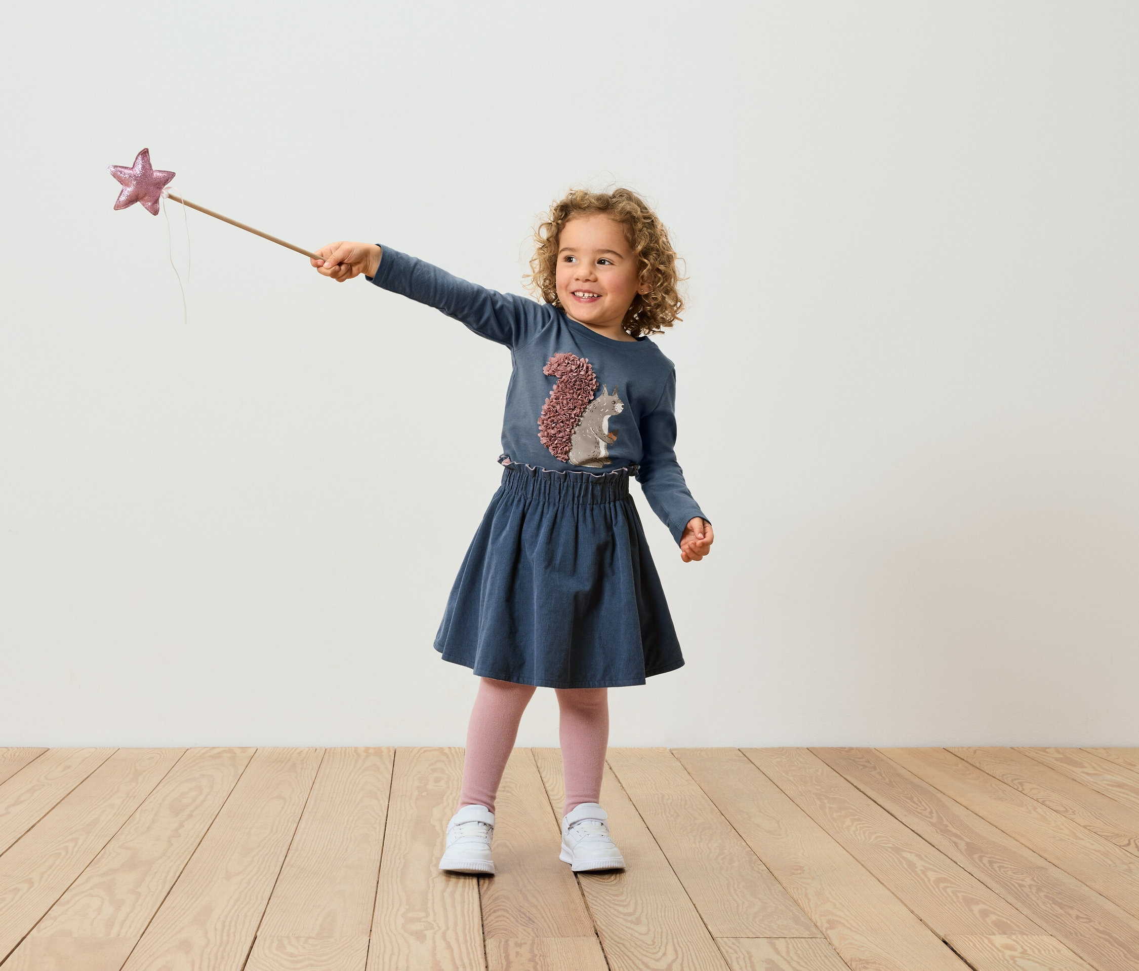 Ein Mädchen mit lockigen Haaren in einem blauen Kleid hält einen Zauberstab mit einem Stern.