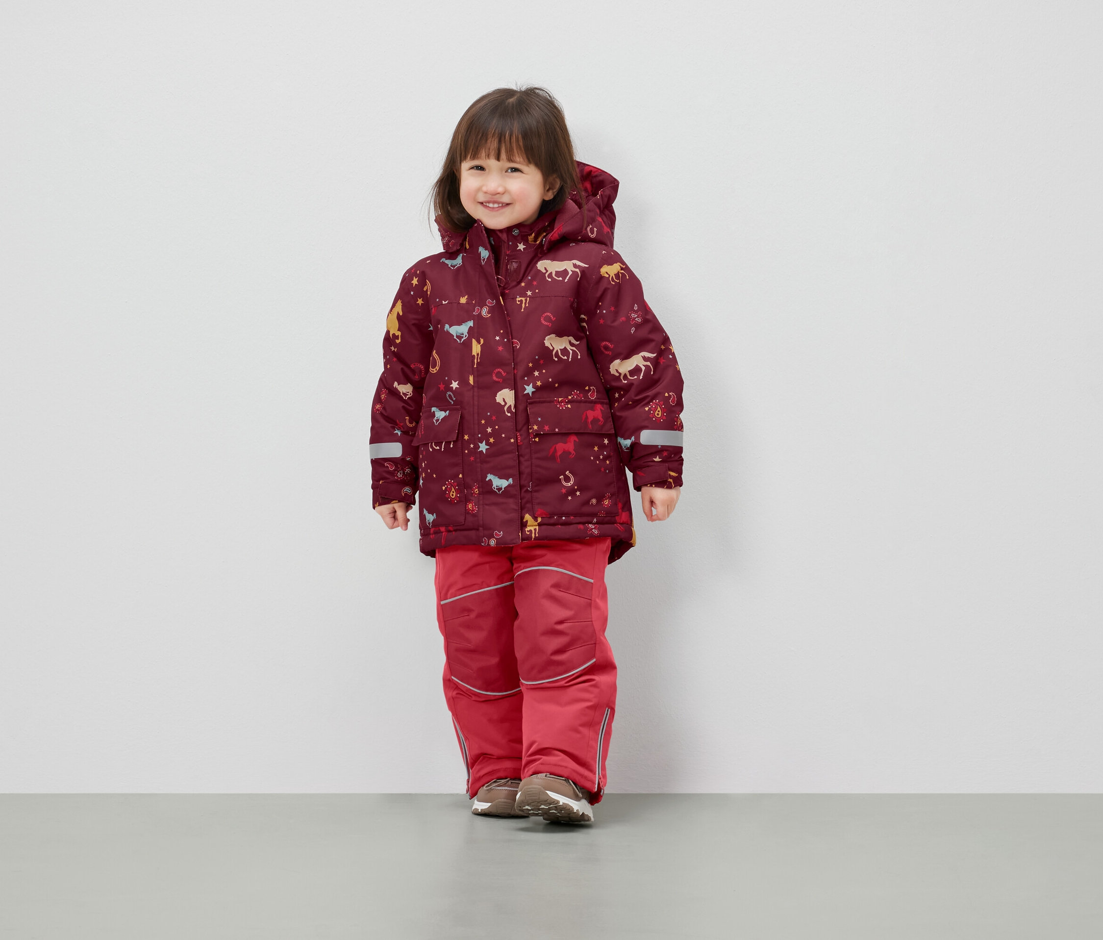 Ein Mädchen steht und trägt eine Winterjacke mit Pferde-Print und eine rote Skihose.