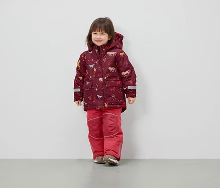 Ein Mädchen steht und trägt eine Winterjacke mit Pferde-Print und eine rote Skihose.