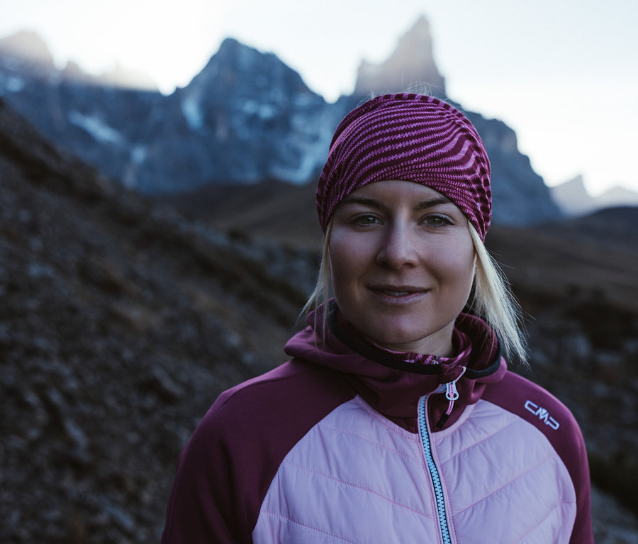Blonde Frau mit gestreiftem Stirnband trägt eine beerefarbene CMP Damen-Hybridjacke vor einer Bergkulisse.