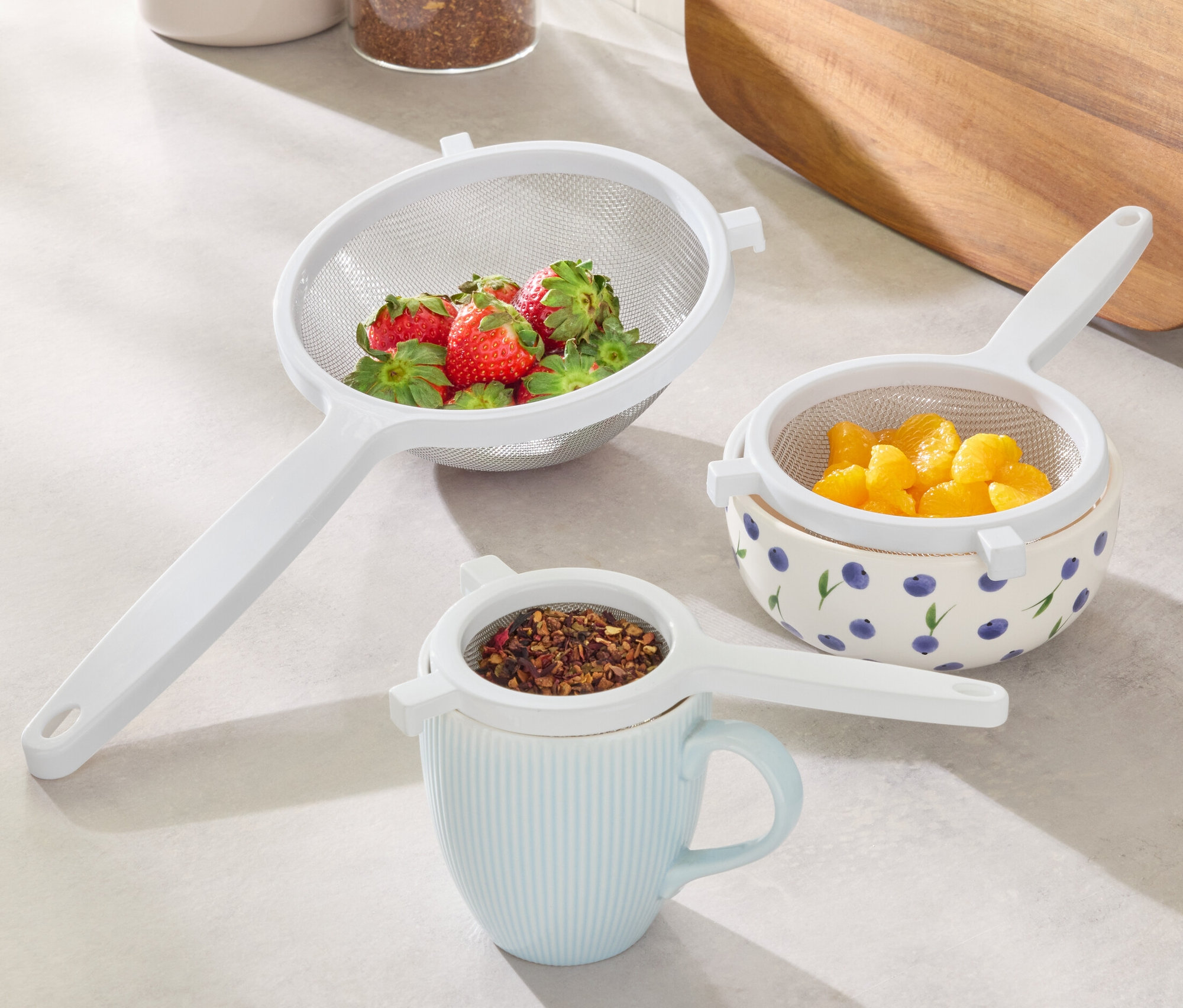 Sieb-Set: Ein Sieb mit Erdbeeren, ein Sieb mit Orangen in einer Schüssel mit Blaubeeren, ein Sieb mit Tee auf einer Tasse.