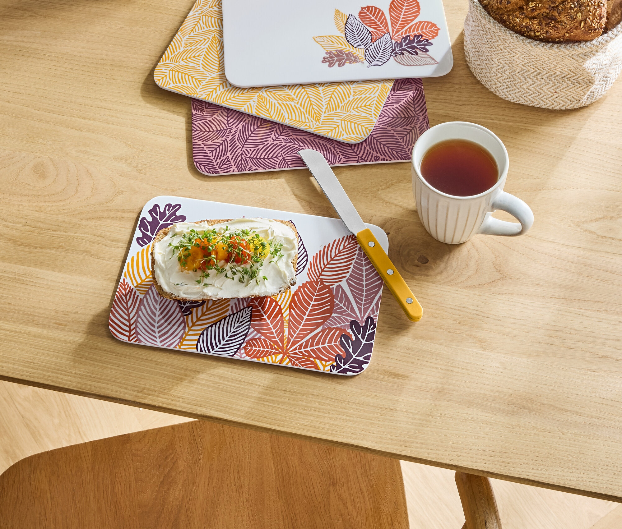 Auf einem Tisch liegen drei Bretter mit Blättermotiven, eines davon mit Brot mit Käse und Gemüse, daneben eine Tasse Tee und ein Brotkorb.