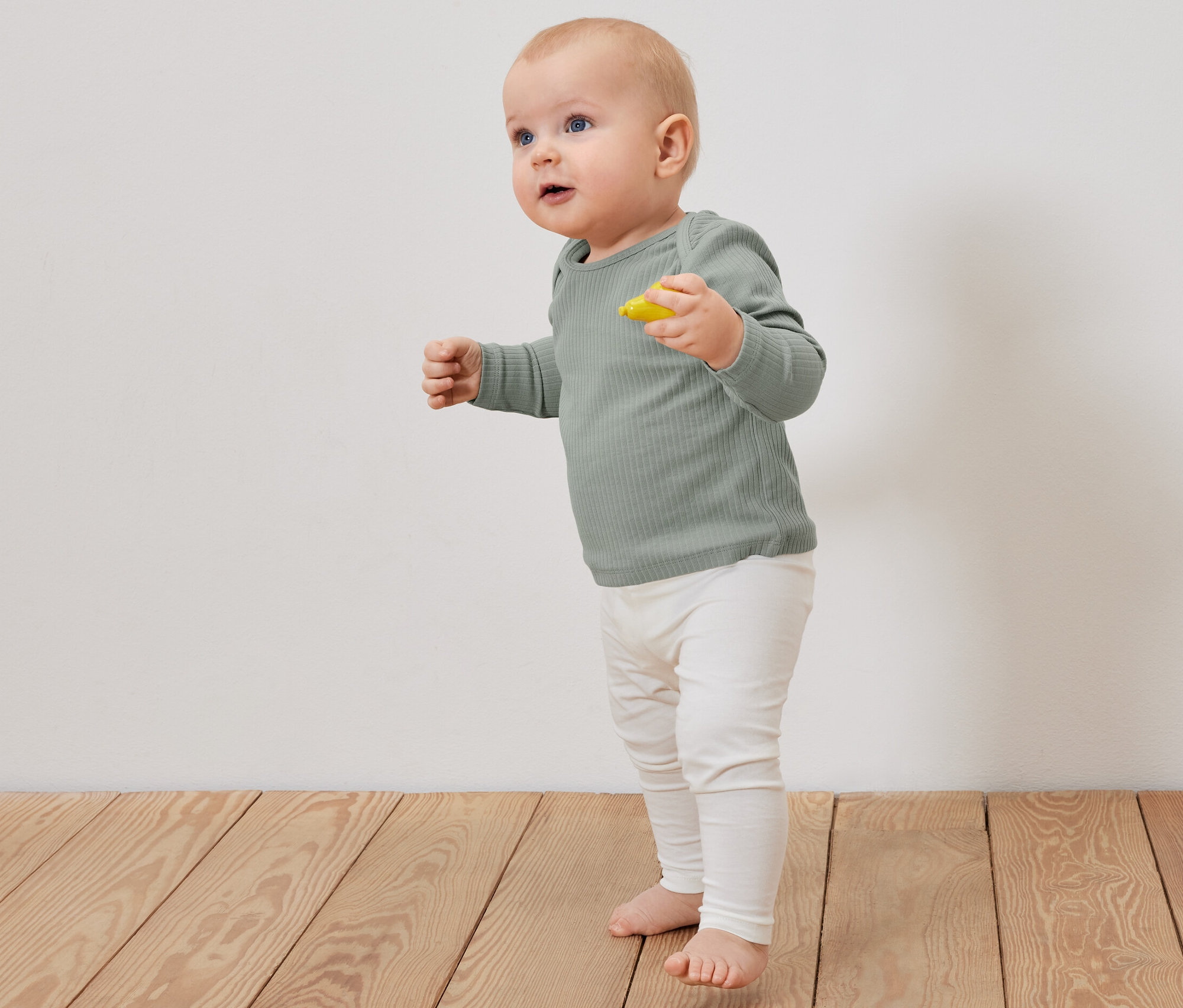 Ein Baby steht auf einem Holzboden und trägt ein hellgrünes geripptes Langarmshirt und beige Leggings.