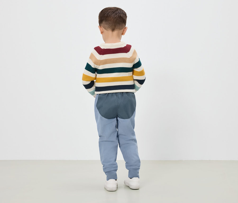 Junge steht mit dem Rücken zur Kamera, trägt einen gestreiften Pullover, blaue Hosen und weiße Turnschuhe.