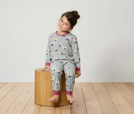 Mädchen sitzt auf einem Holzhocker und trägt einen grauen Schlafanzug mit Ballerinas.