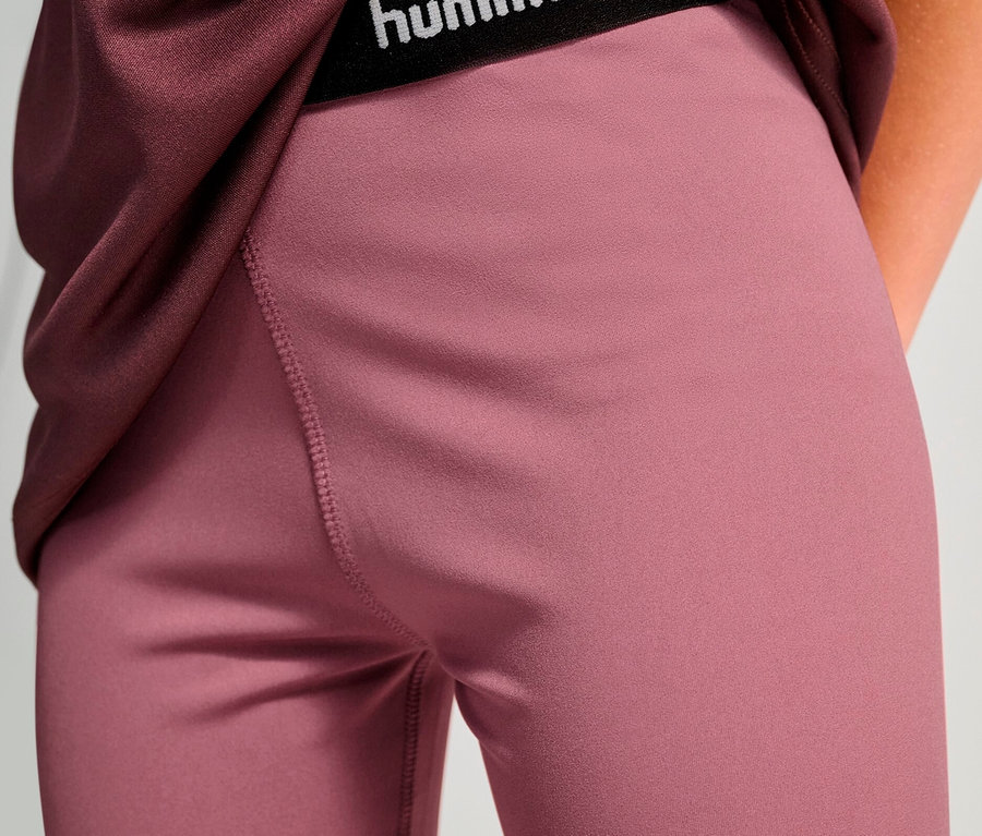 Nahaufnahme einer Person, die ein weinrotes Oberteil und pinke HUMMEL HMLJR Pulse MW Short Tights mit schwarzem "hummel"-Logobund trägt.