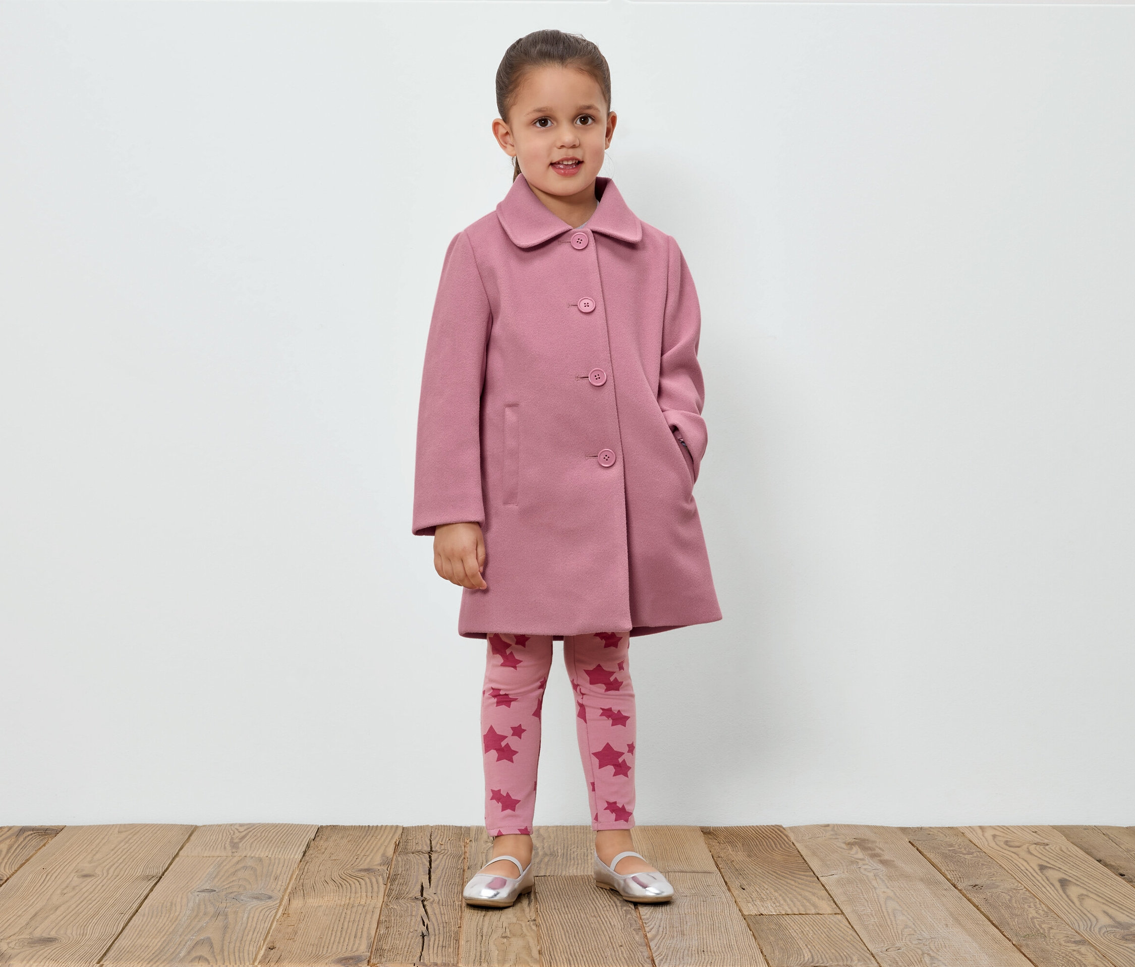 Mädchen steht vor einer weißen Wand und trägt einen rosa Mantel, Leggings mit Sternen und silberne Schuhe.