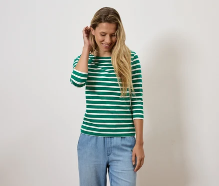 Blonde Frau lächelt und richtet ihr Haar, trägt ein grün-weiß gestreiftes Shirt und blaue Jeans.