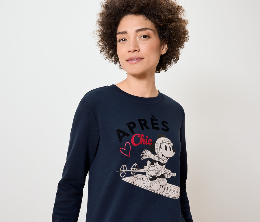 Eine Frau trägt einen dunkelblauen Pullover mit einem Aufdruck von Mickey Mouse auf einem Snowboard.